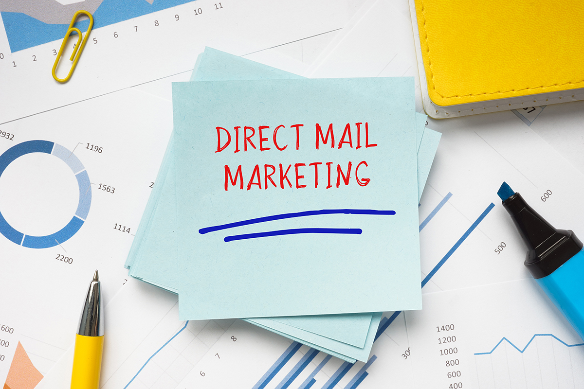 An image of a blue sticky note that says “direct mail marketing” in red. Other office supplies and papers surround the message.