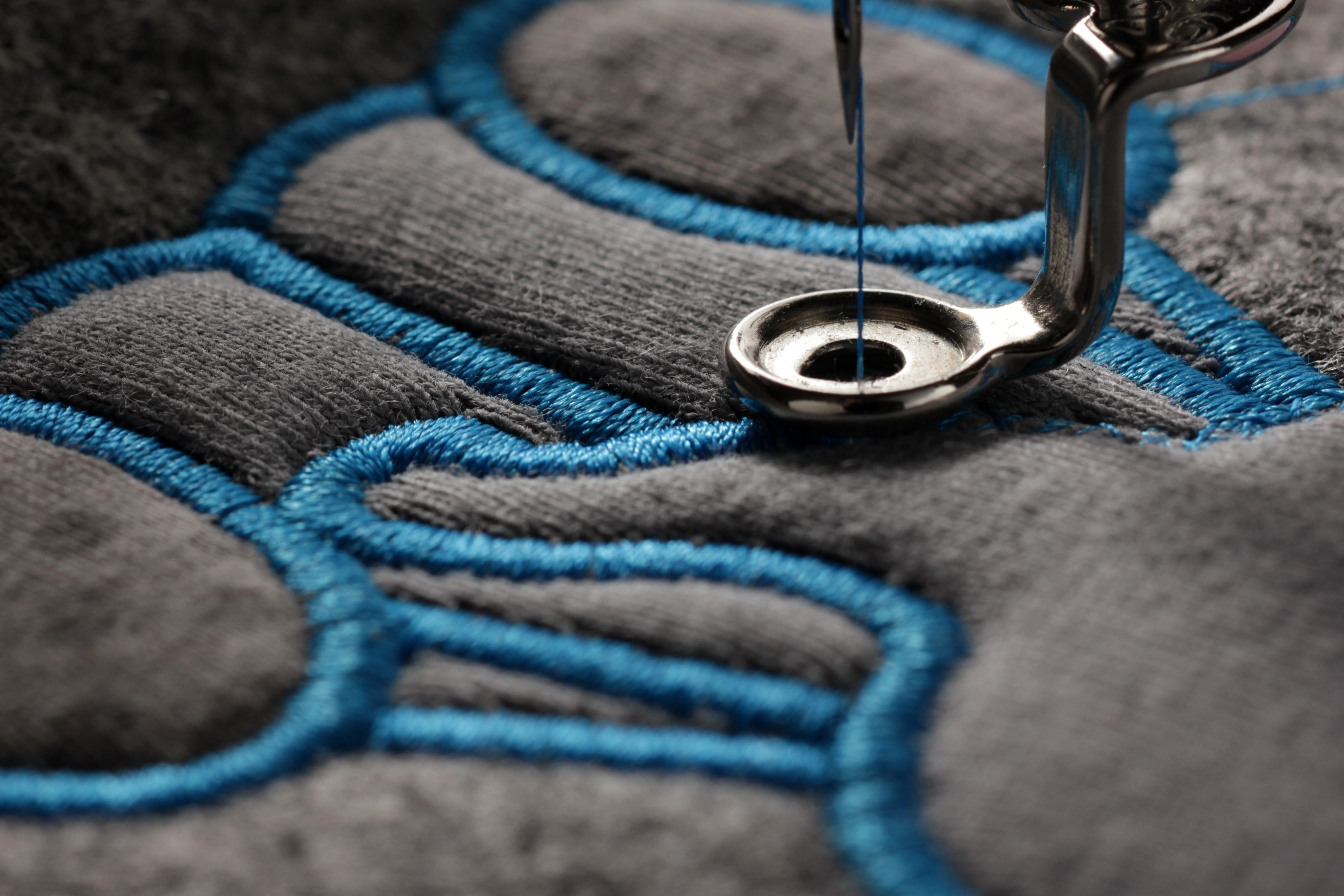 A sewing machine stitches a blue logo into a gray fabric