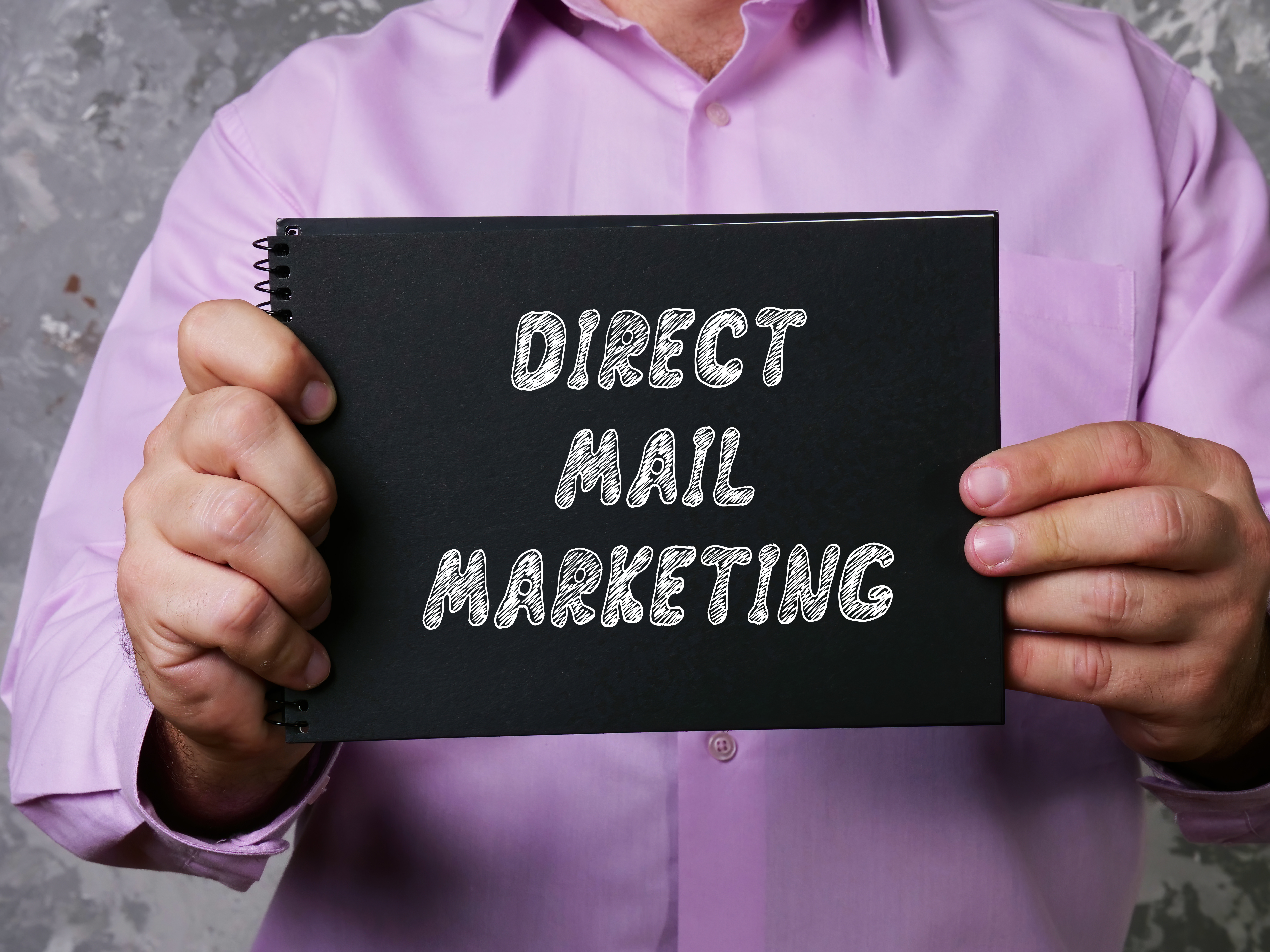 an image of a person holding up a black piece of paper that says “direct mail marketing” in white font.