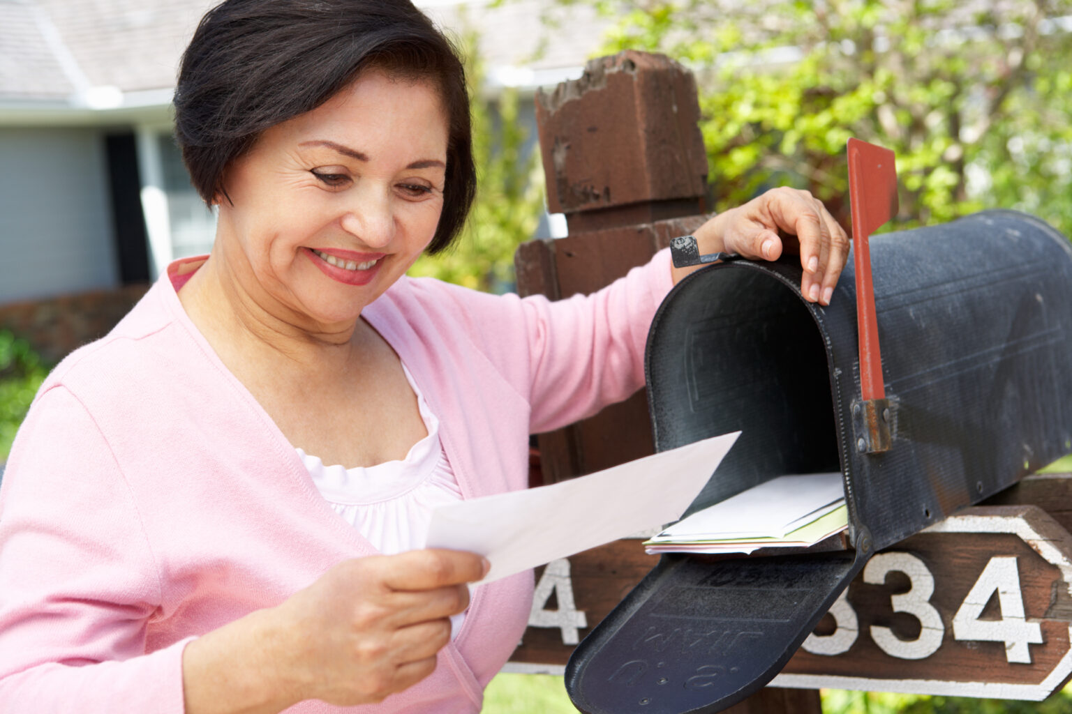 A woman reads her mail and smiles, standing in front of her open mailbox
